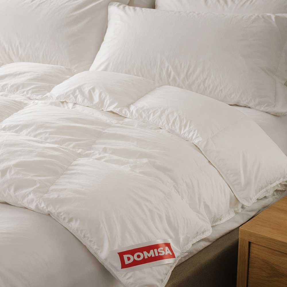 Pilotă Ovia 200x220 cm pe pat cu perne albe, umplutură naturală din puf și pene de gâscă, design elegant și aerisit.